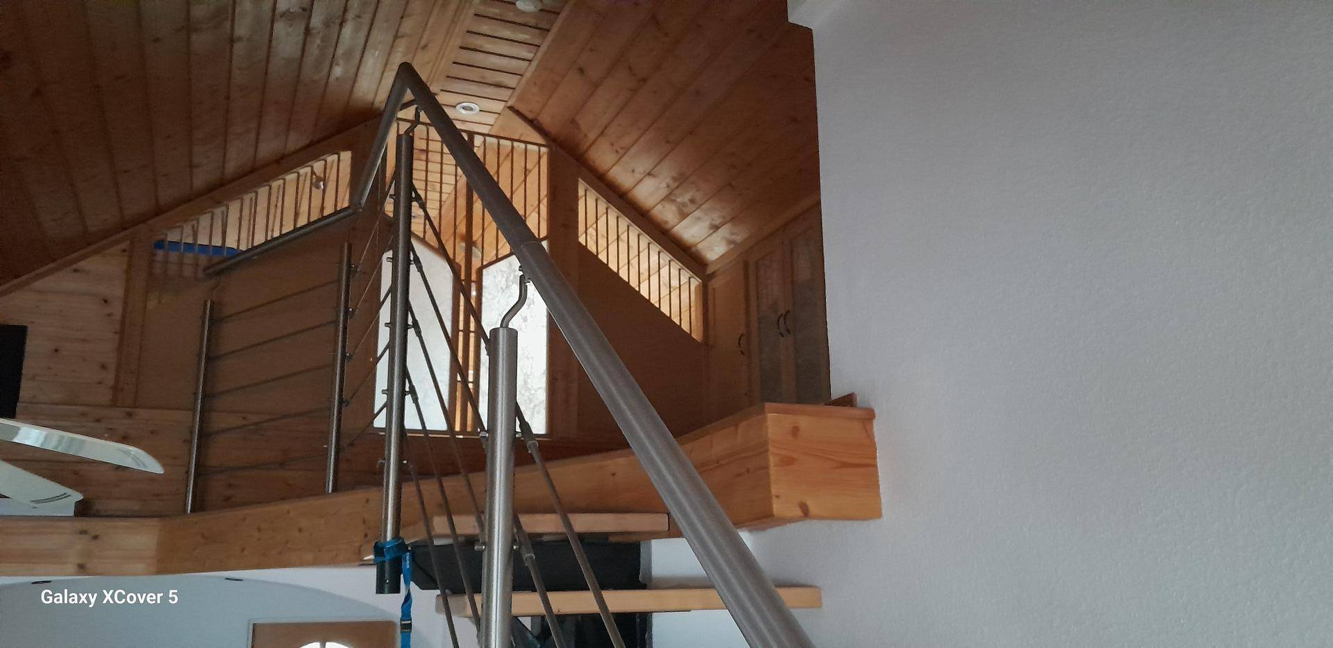 Detailaufnahme einer Holzdecke und einer Treppe mit Metallgeländer, die zum Obergeschoss führt.