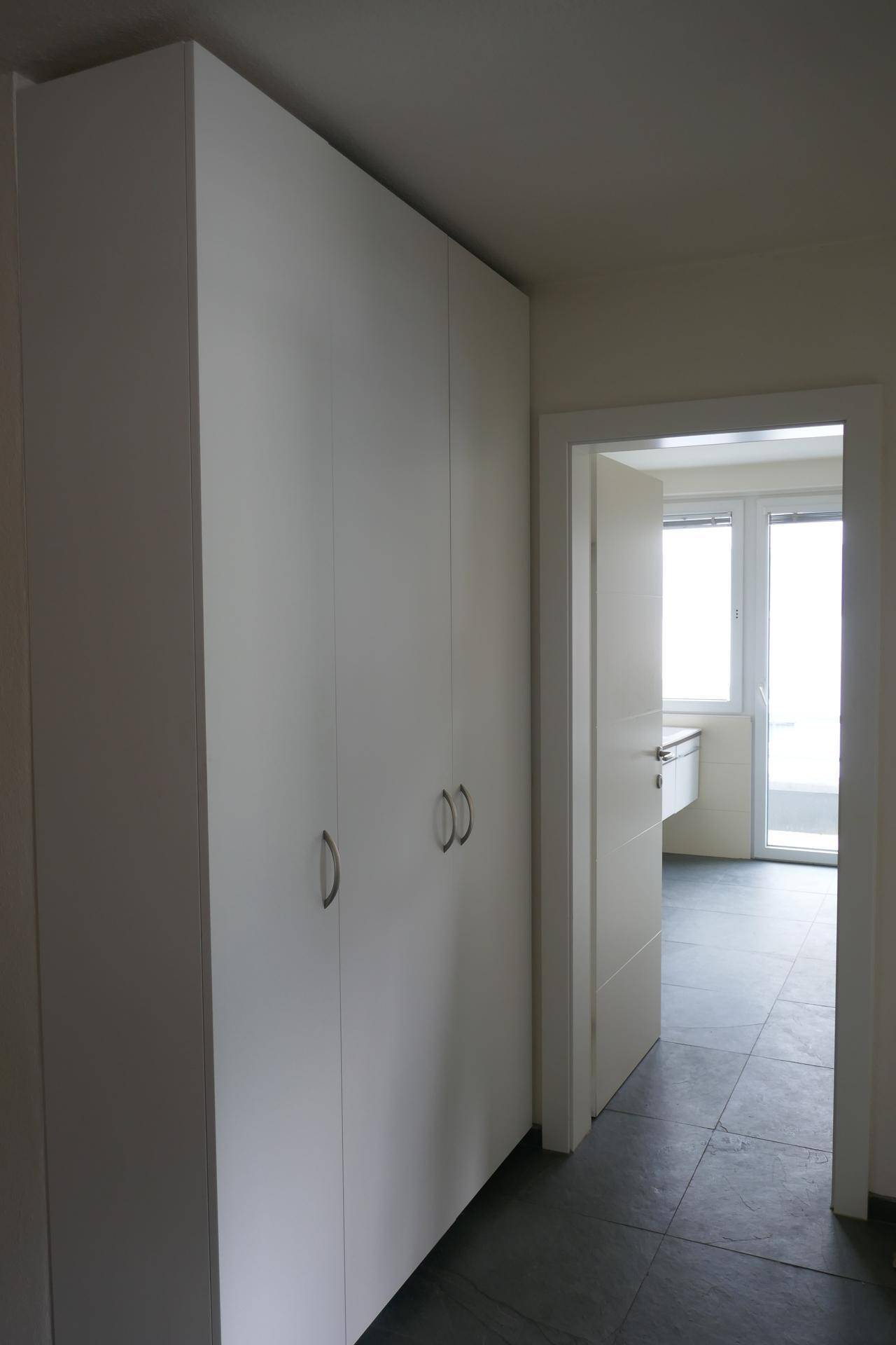 Flur mit weißen Einbauschränken und Blick in ein helles Badezimmer mit Fenster.