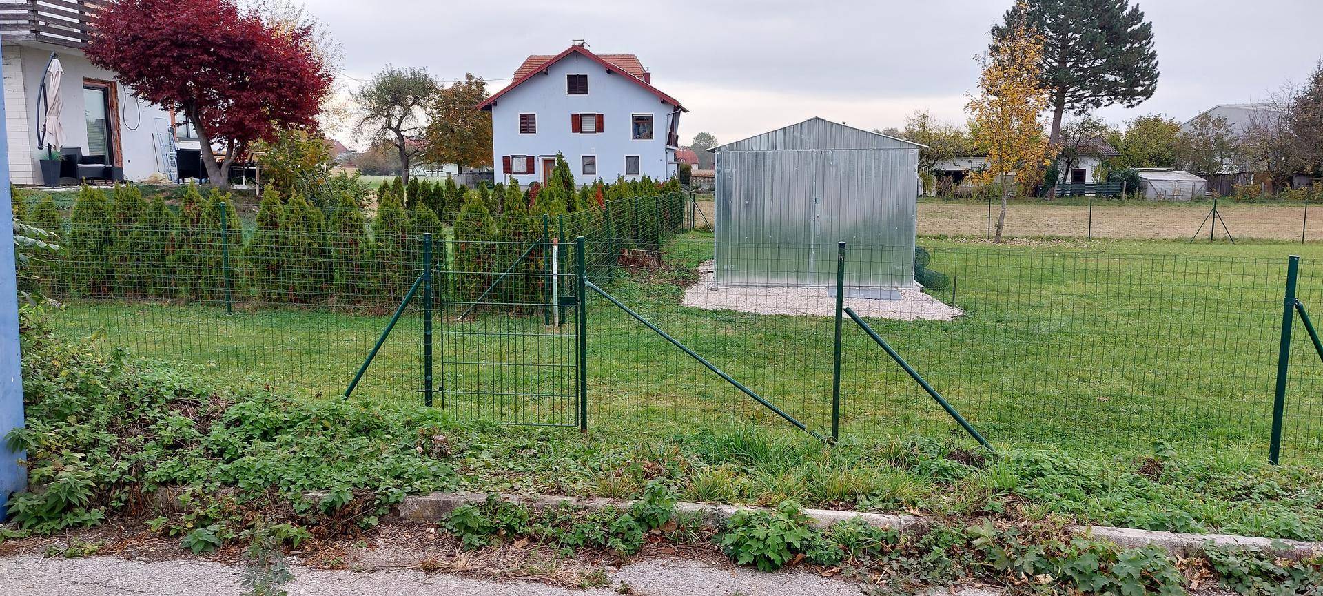 Eingezäunter Gartenbereich mit Rasen, einem Metallschuppen und Wohnhäusern im Hintergrund an einem sonnigen Tag.