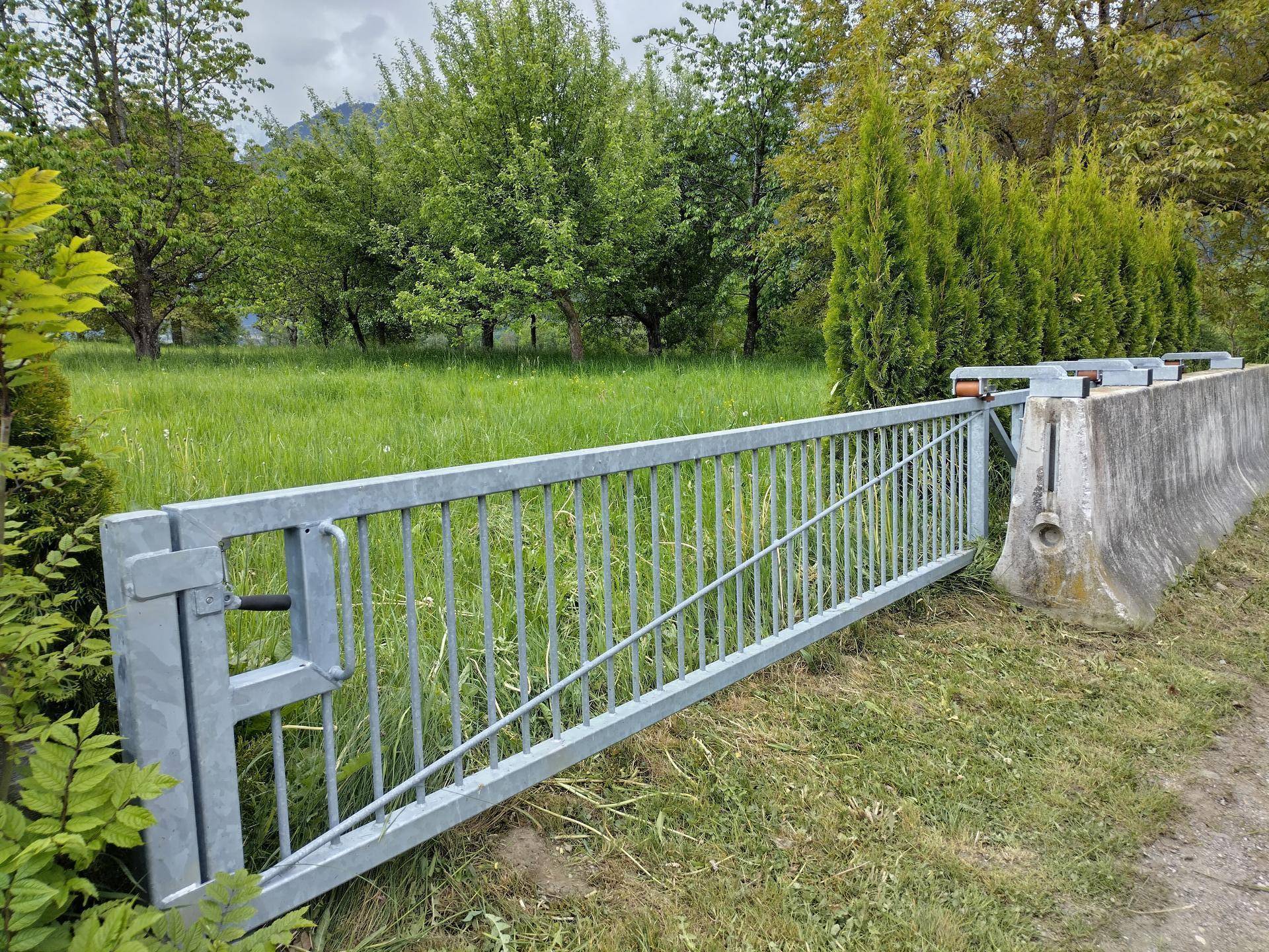 Metalltor mit Betonbegrenzung und Hecke, umgeben von üppigem Grün und Bäumen.