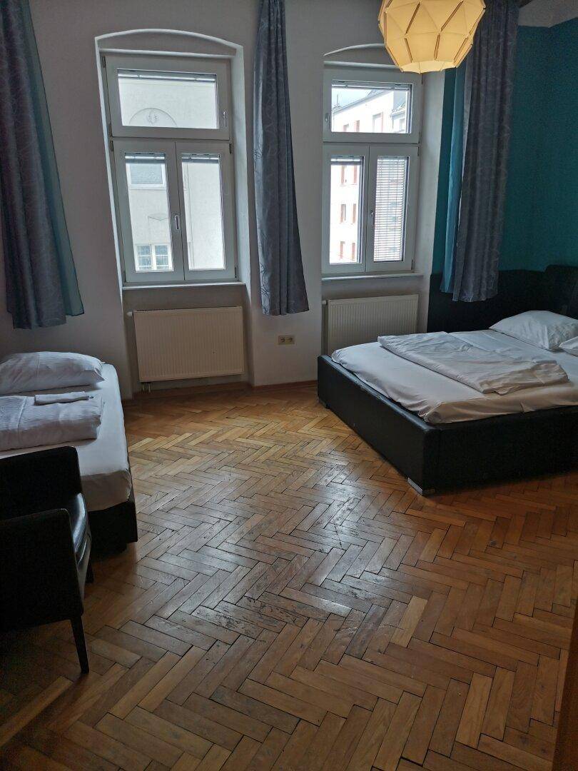 Helles Schlafzimmer mit zwei Betten, Fischgrätparkett und hohen Fenstern.