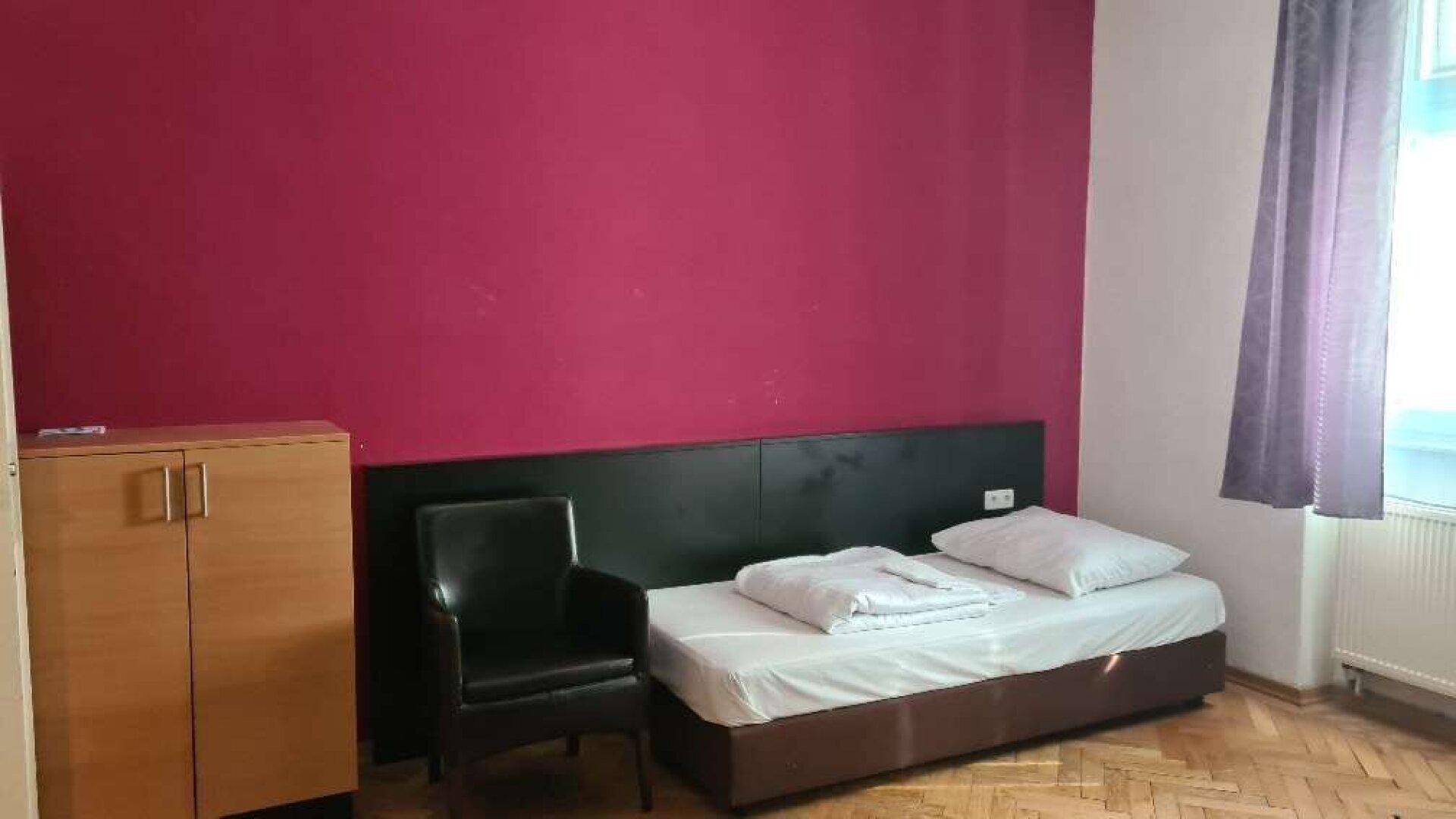Schlafzimmer mit Einzelbett, Sessel und auffälliger magentafarbener Wandgestaltung.