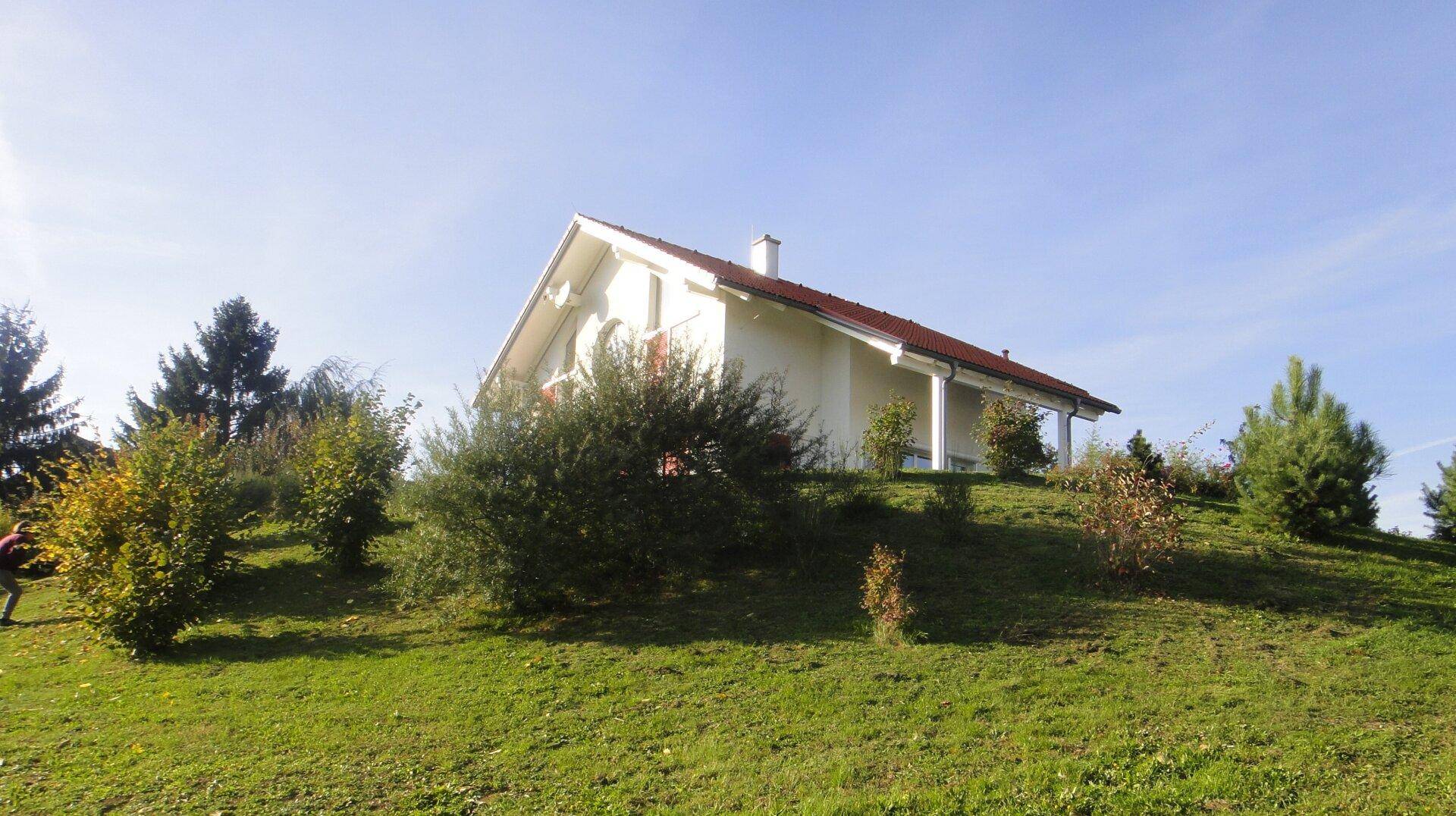 Modernes Einfamilienhaus auf einem Hügel, umgeben von grüner Landschaft und Sträuchern unter blauem Himmel.