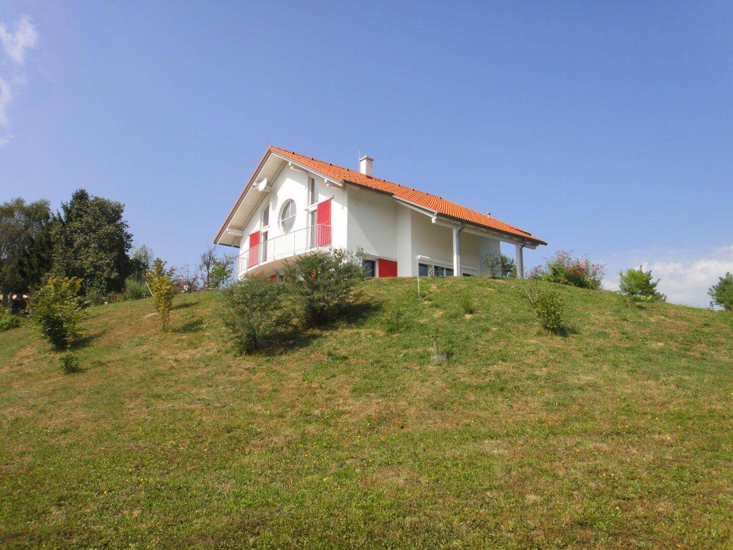 Freistehendes Haus mit weißer Fassade und rotem Ziegeldach auf einem sonnigen Hügelgrundstück mit Weitblick.