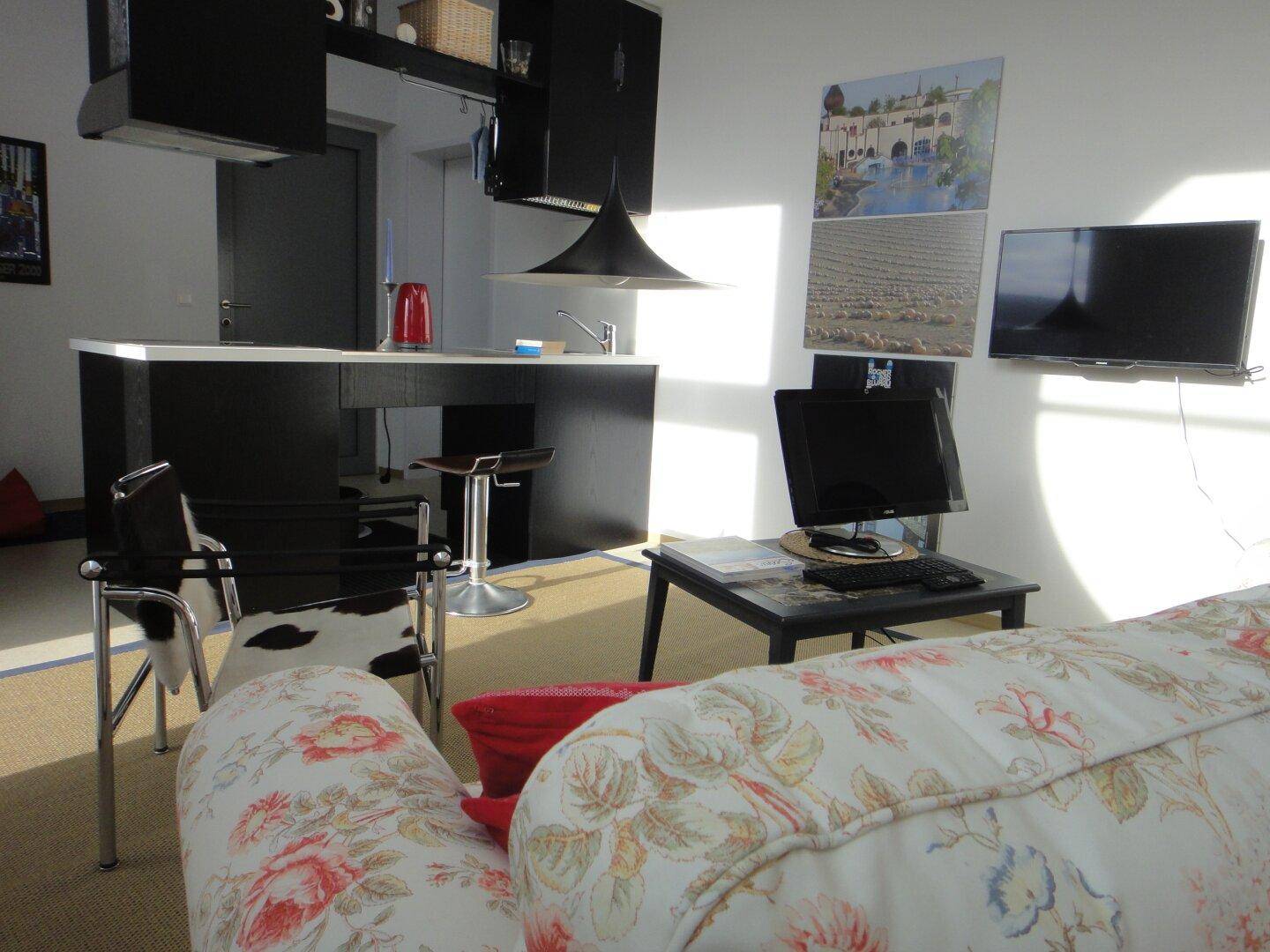 Helles Wohnzimmer mit floralem Sofa, Fernseher und einer modernen Küchenzeile im Hintergrund.