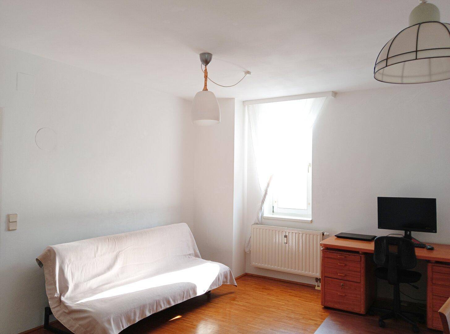 Helles Zimmer mit Holzboden, Sofa und Schreibtisch, ideal als Schlaf- oder Arbeitszimmer.