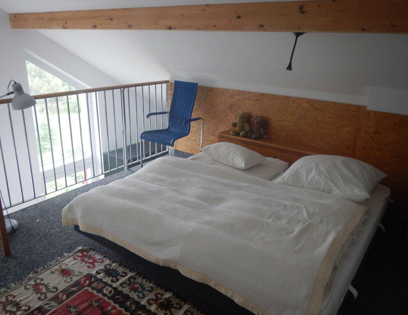 Helles Schlafzimmer im Dachgeschoss mit Holzbalken, Teppichboden und Blick durch die Fenster.