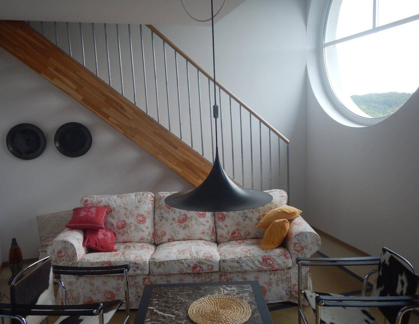 Wohnzimmer mit floralem Sofa, Treppe und großem runden Fenster, das viel Licht hereinlässt.