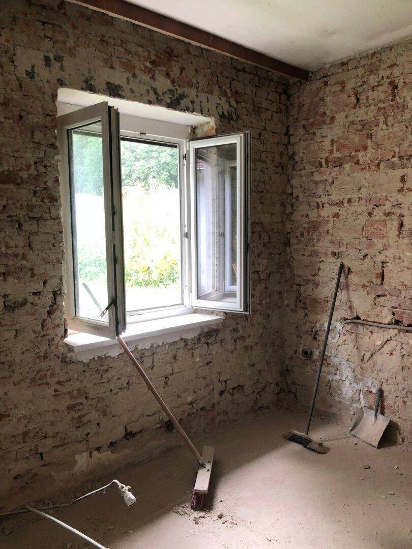 Heller Raum im Rohbauzustand mit freigelegten Ziegelwänden und geöffnetem Fenster zum Garten.