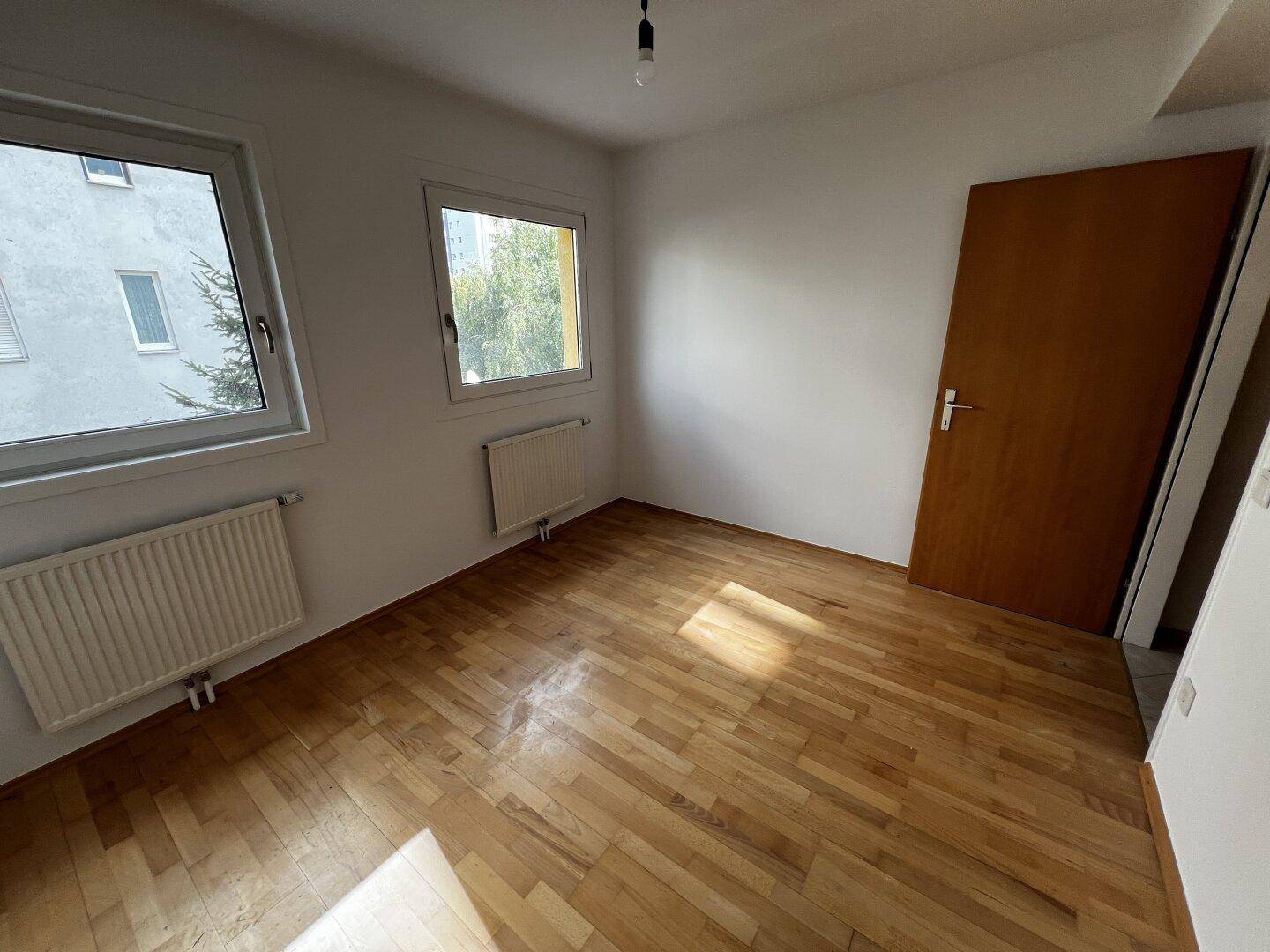 Helles Schlafzimmer mit Parkettboden, zwei Fenstern und einem Heizkörper, bereit zum Einrichten.