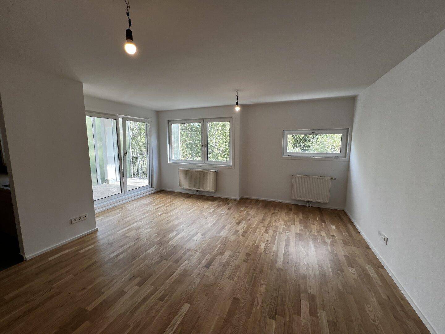 Geräumiges Wohnzimmer mit hellem Parkettboden, mehreren Fenstern und einem Balkonzugang.