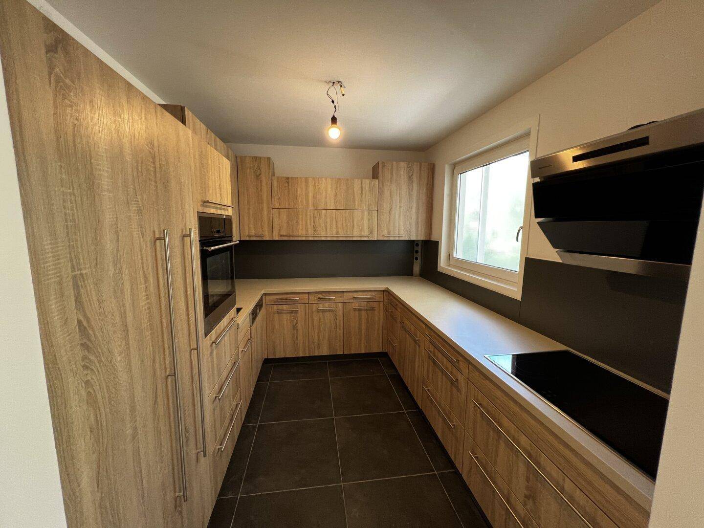 Moderne Einbauküche mit Holzfronten, dunklen Fliesen, integrierten Geräten und einem Fenster.