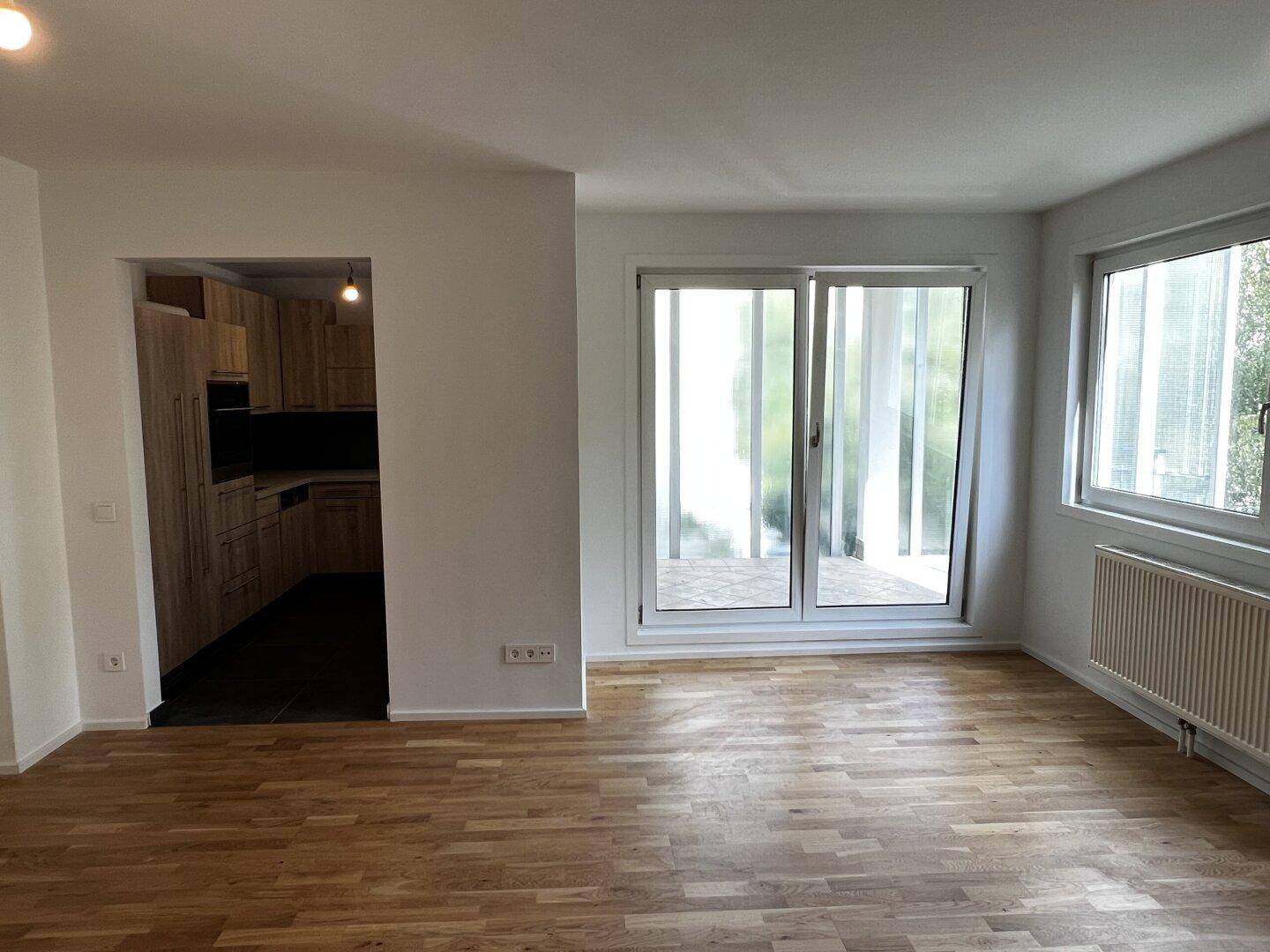Lichtdurchflutetes Wohnzimmer mit Holzboden, Balkonzugang und Blick auf die angrenzende Küche.