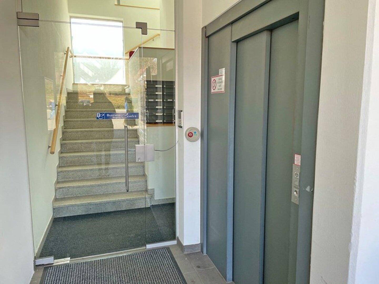 Eingangsbereich mit Aufzug, Treppe mit Glasgeländer und Zugang zu weiteren Etagen.
