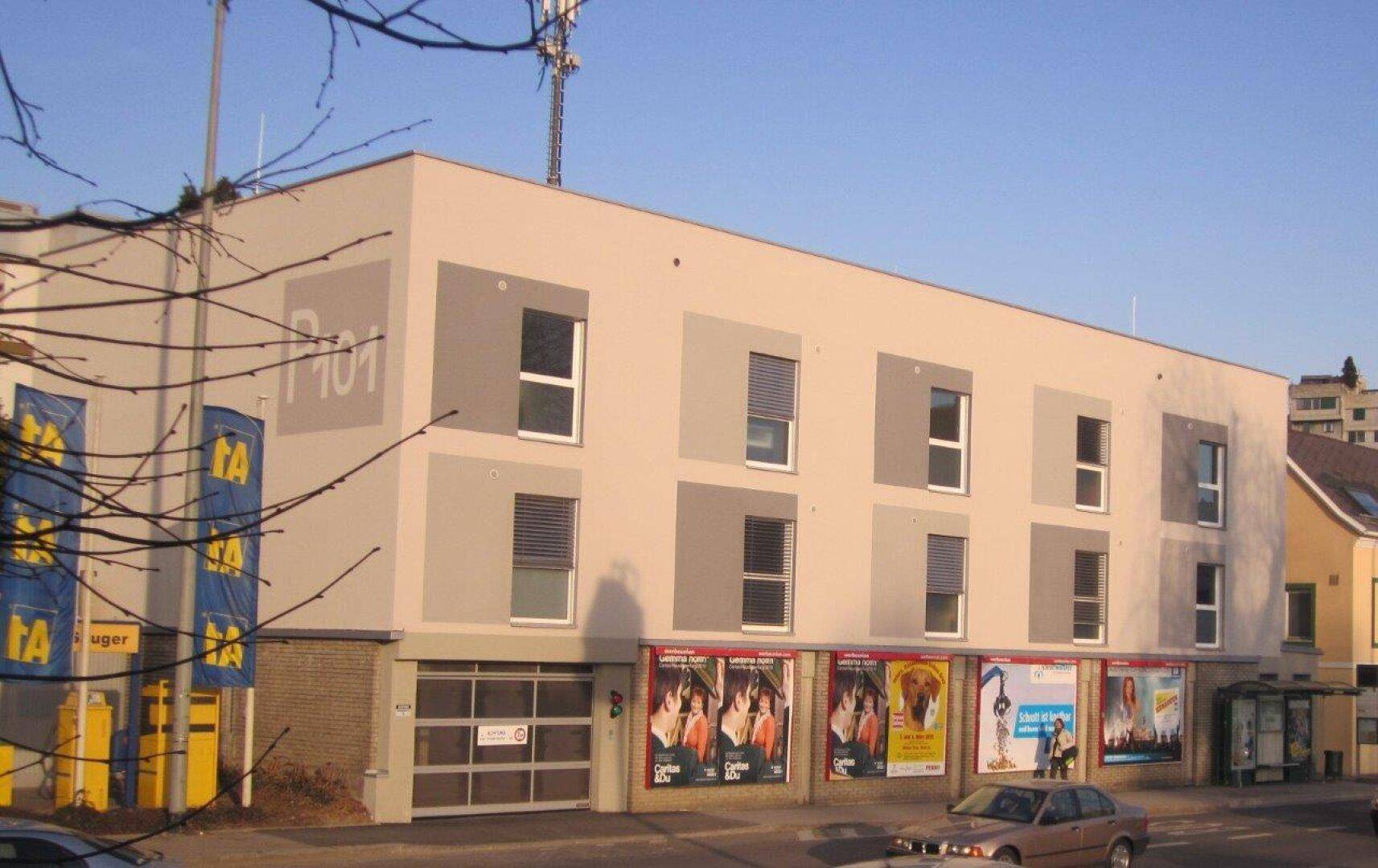 Modernes Gebäude mit beiger Fassade, großen Fenstern und Geschäften im Erdgeschoss an einer Straße.