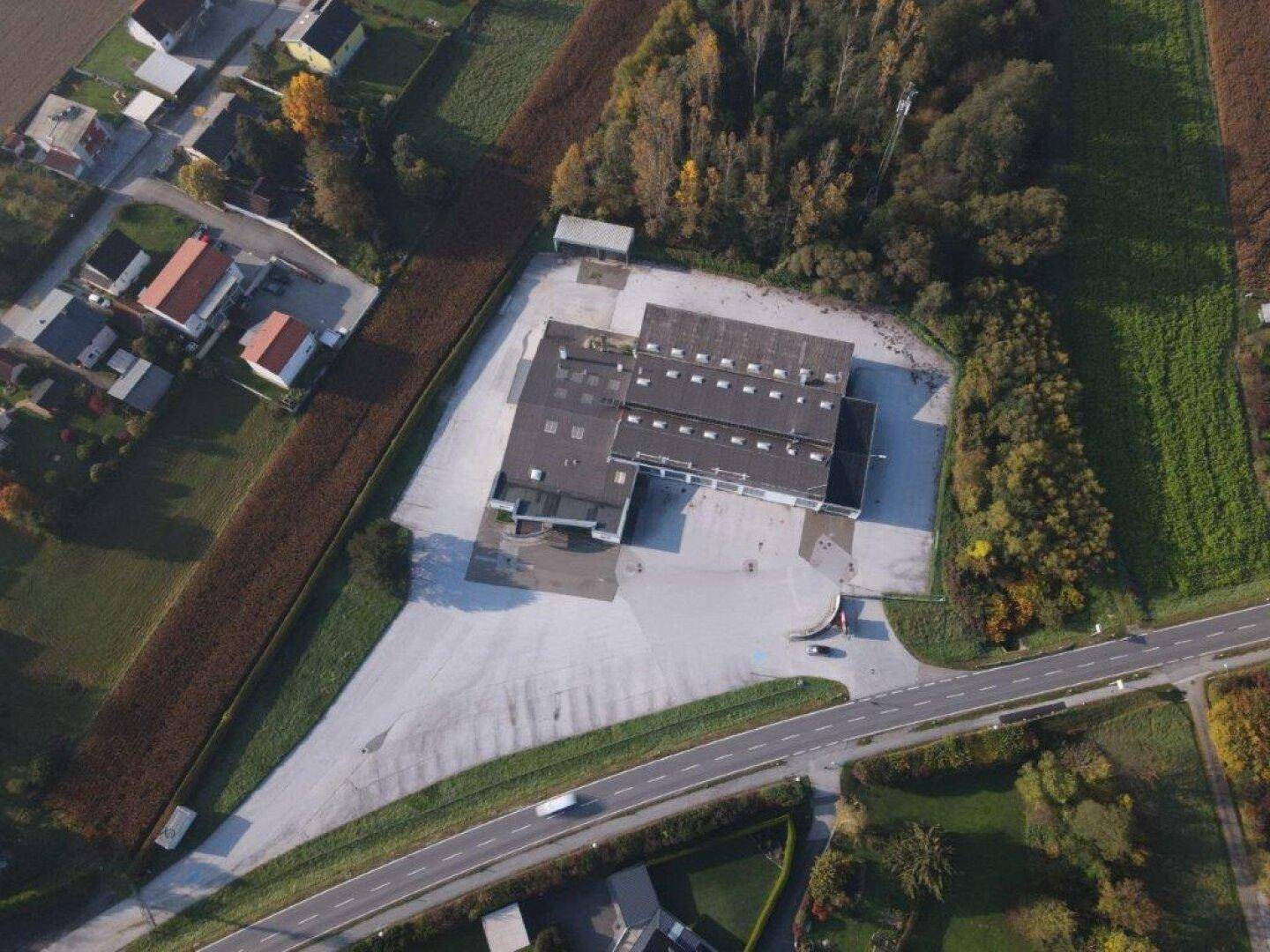Luftaufnahme eines Gewerbeobjekts mit großem Parkplatz, umgeben von Feldern und einer Straße.