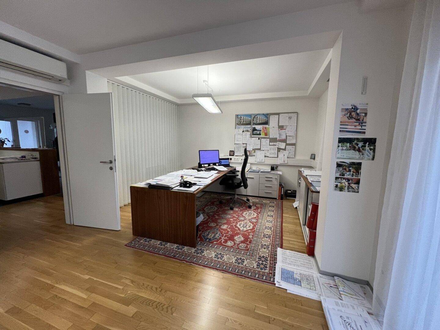 Geräumiges Büro mit Holzschreibtisch, Teppich und einem großen Fenster mit Jalousien.