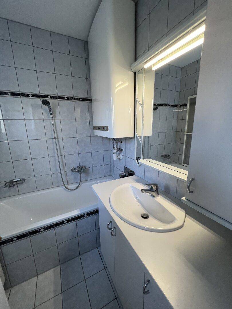Badezimmer mit hellgrauen Fliesen, Badewanne mit Duschfunktion und Spiegelschrank.
