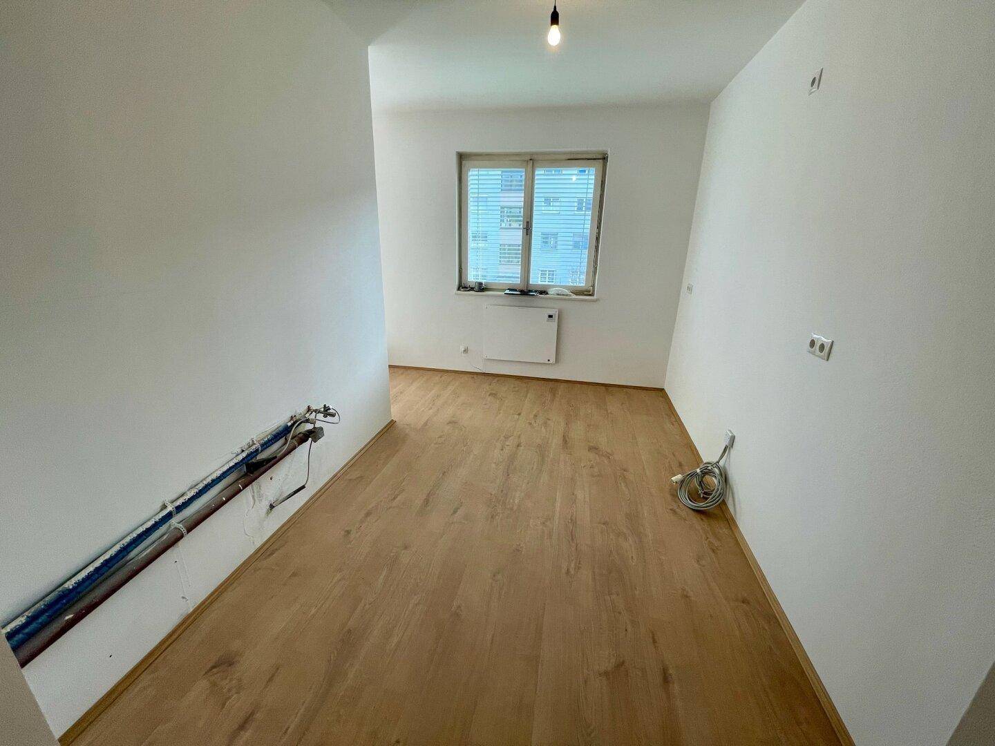 Leerer Raum mit Holzfußboden und vorbereiteten Anschlüssen für eine Küche.