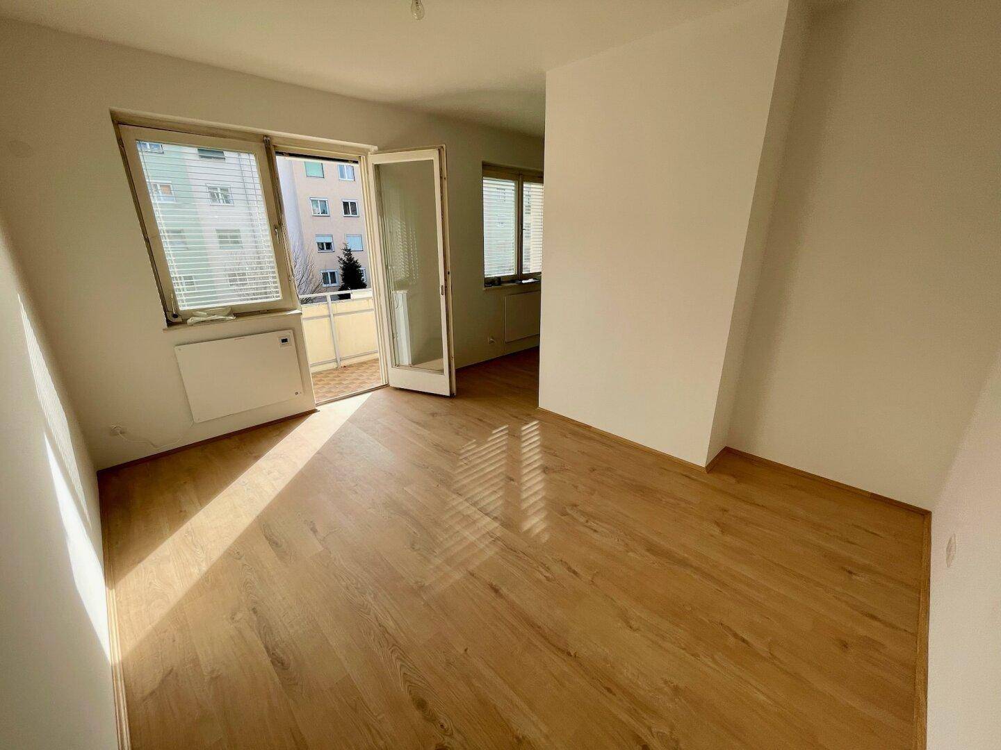 Lichtdurchfluteter Raum mit Holzfußboden, großem Fenster und Zugang zum Balkon.