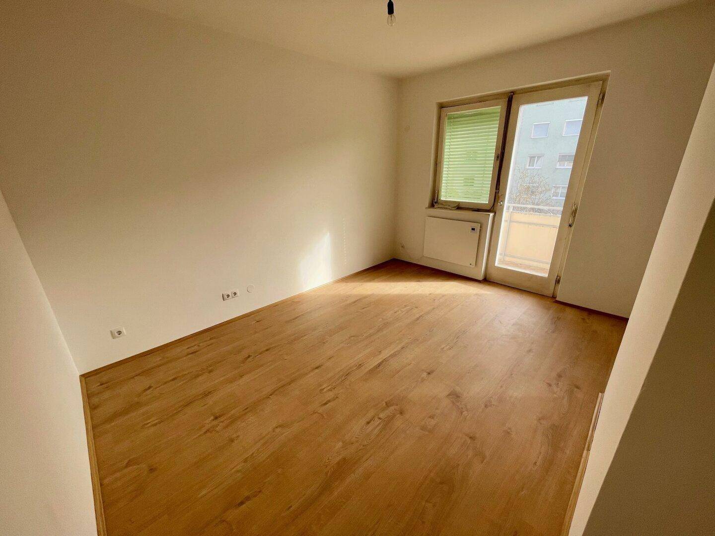 Lichtdurchfluteter Raum mit Holzfußboden und Zugang zu einem sonnigen Balkon.