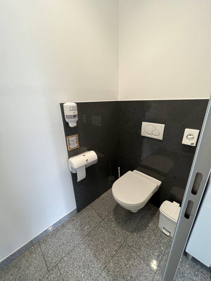 Saubere Toilette mit wandhängendem WC und dunkler Rückwand, in einem gefliesten und gut gepflegten Bereich.