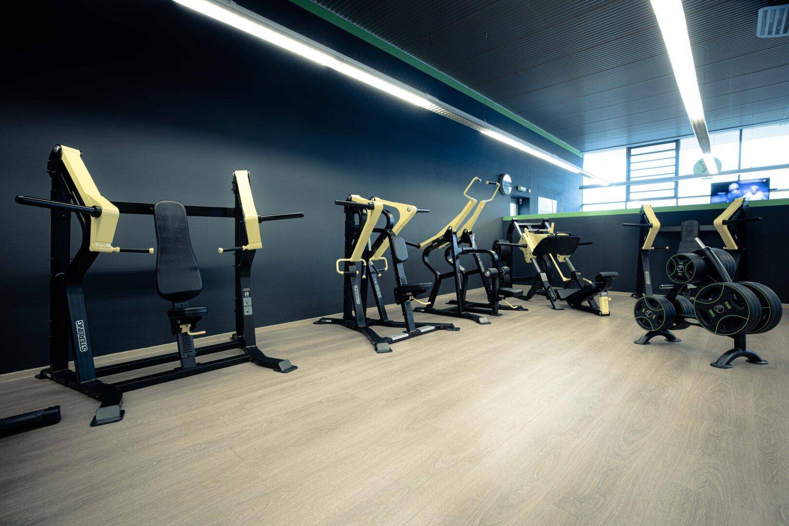 Bereich mit Krafttrainingsgeräten in einem Fitnessstudio, ausgestattet mit dunklen Wänden und hellem Boden.