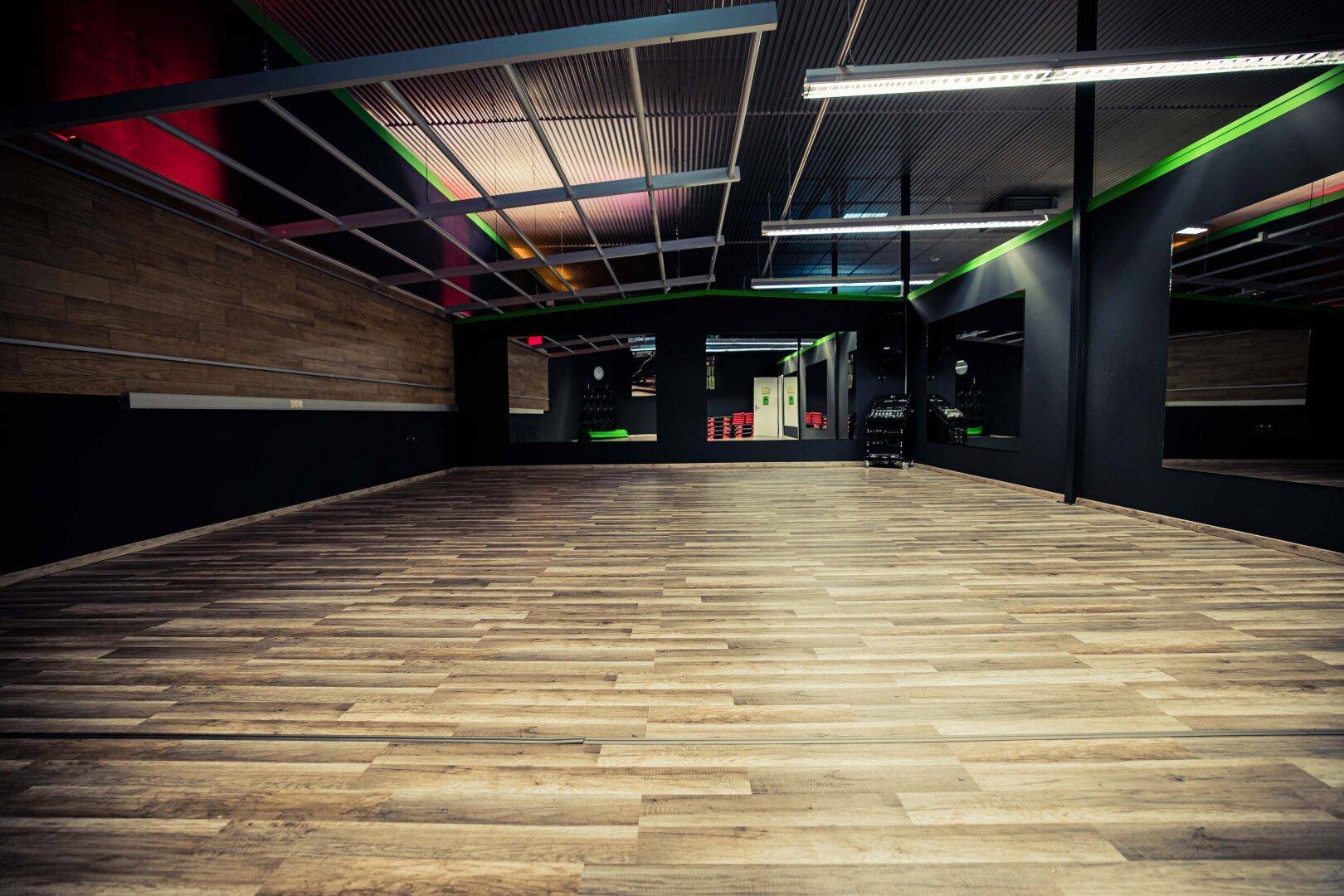 Geräumiger Kursraum mit Holzboden und dunklen Wänden, bereit für verschiedene Fitnessaktivitäten.