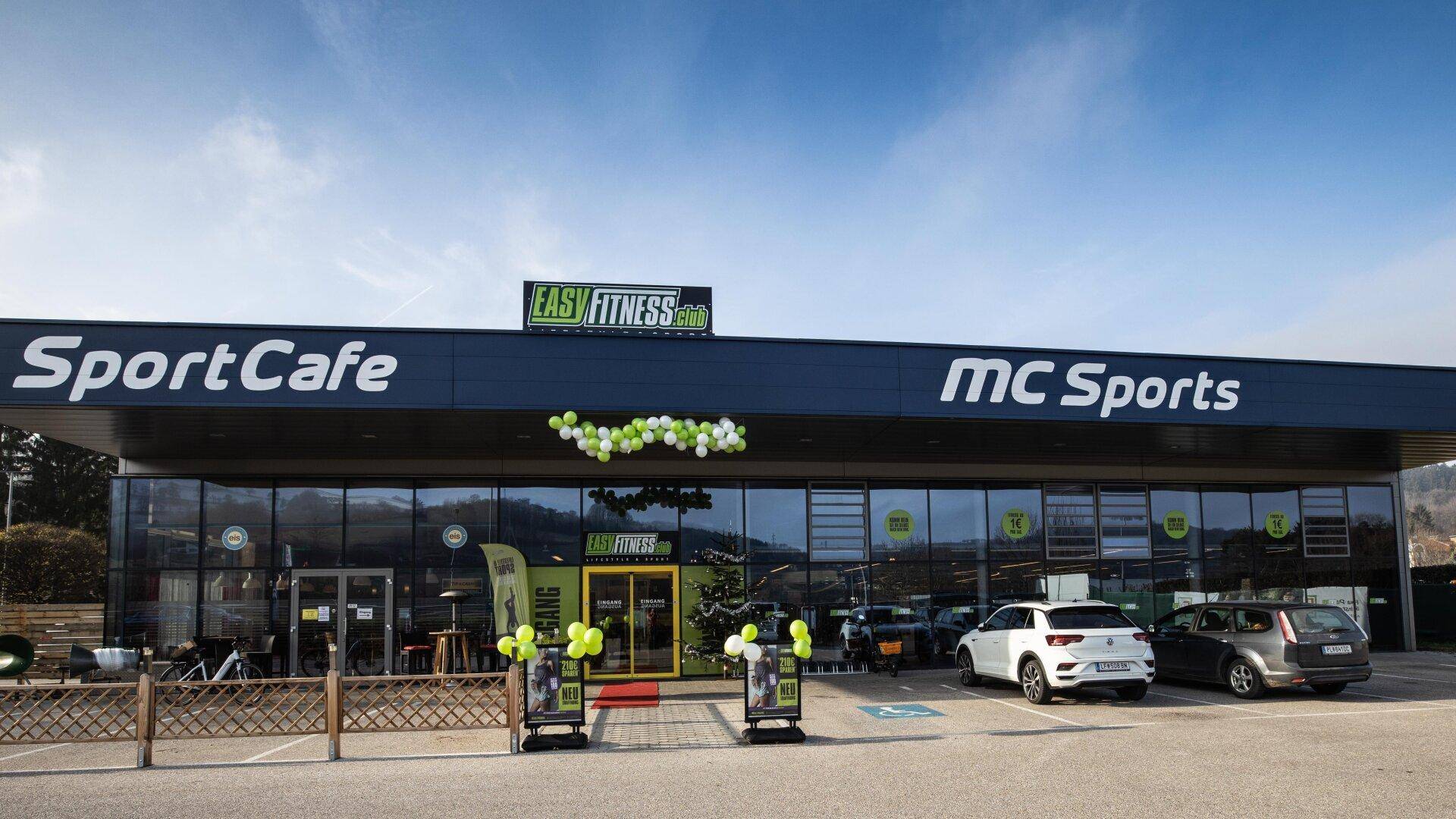 Ansicht des modernen Geschäftsgebäudes mit Fitnessstudio und Café, umgeben von Parkplätzen.