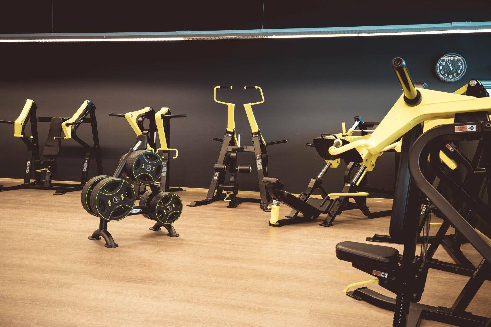 Fitnessbereich mit verschiedenen Krafttrainingsgeräten in Schwarz und Gelb auf hellem Holzboden.