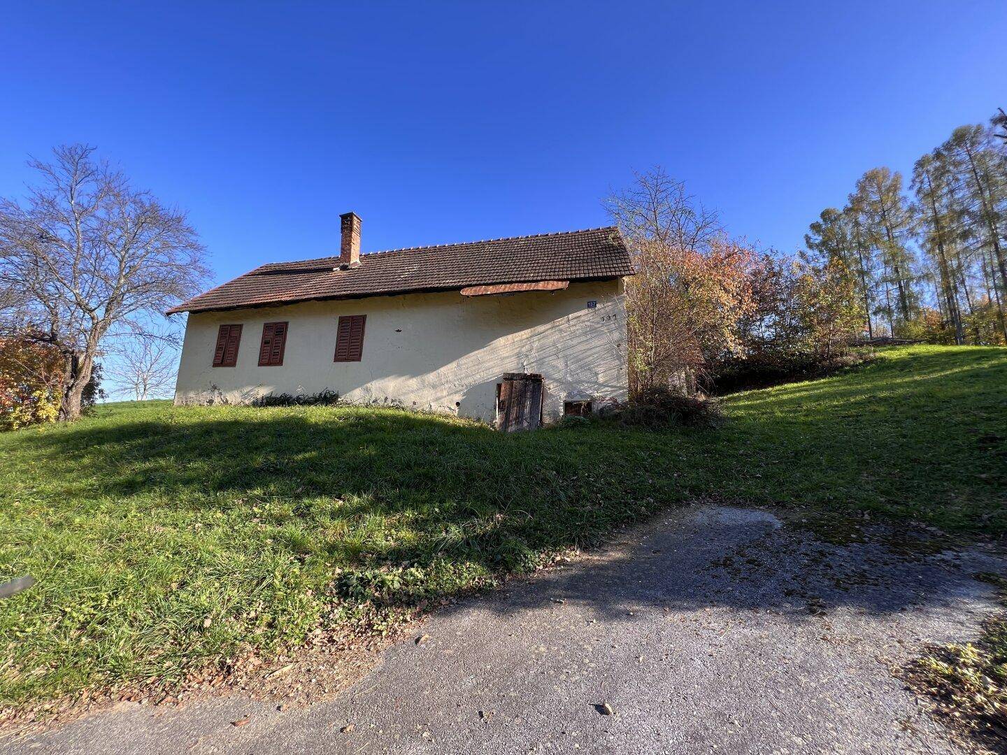 Seitenansicht eines alten Hauses mit Ziegeldach und Fensterläden auf einem sonnigen, grasbewachsenen Hügel.