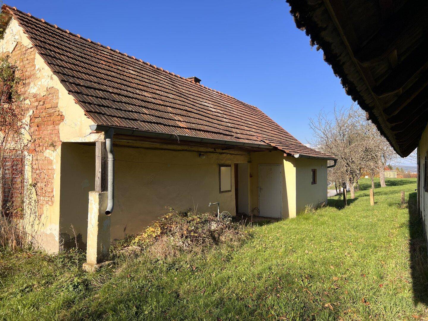 Seitenansicht eines alten Hauses mit überdachtem Eingang und verwitterter Fassade auf einem grasbewachsenen Grundstück.
