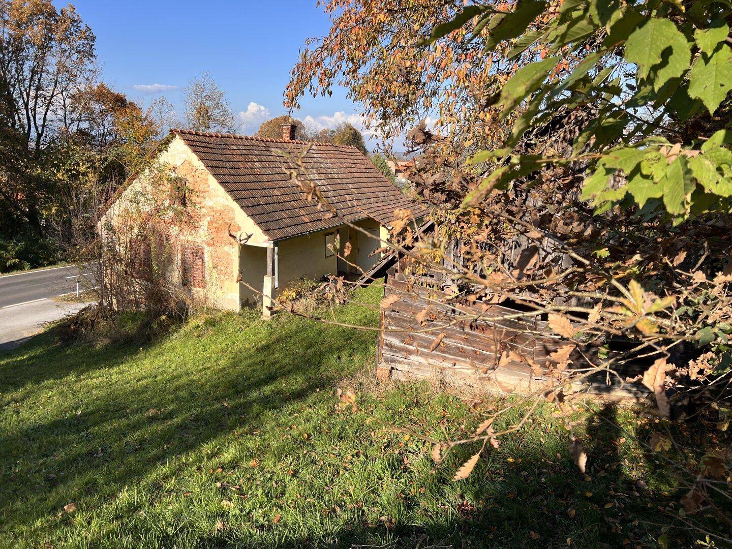 Verfallenes Haus mit Ziegeldach und angrenzendem Holzschuppen auf einem sonnigen Hang.