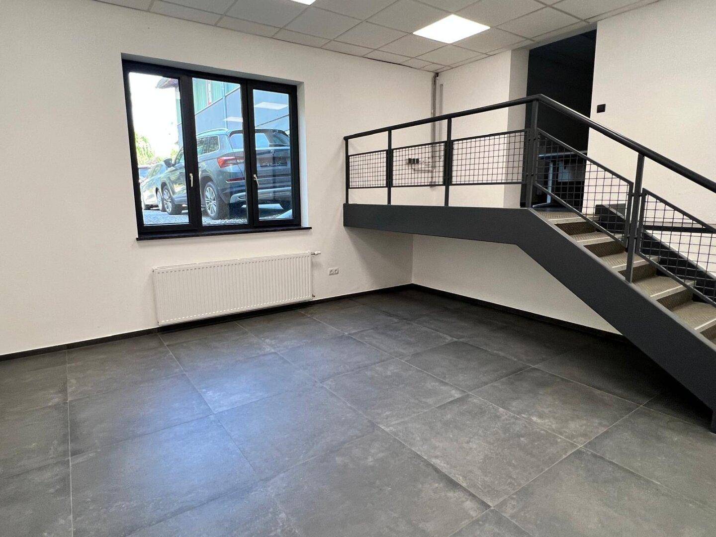 Heller Büroraum mit dunklen Fliesen, einem großen Fenster mit Blick auf parkende Autos und einer schwarzen Treppe.