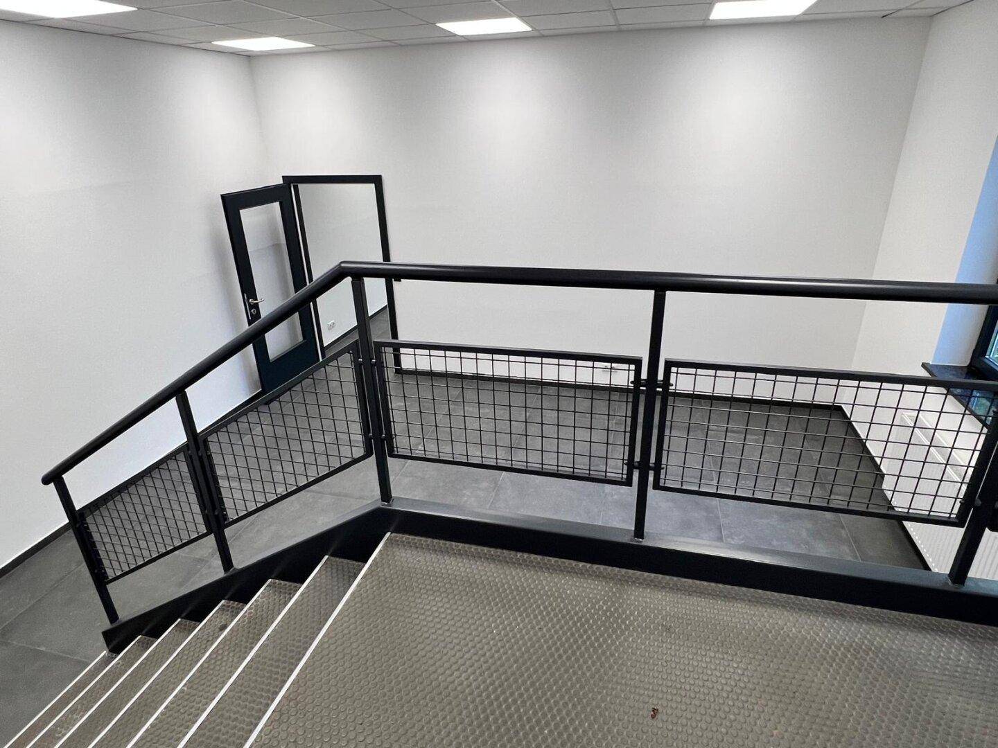 Moderne Treppe im Industriestil mit schwarzen Metallgeländern und dunklem Fliesenboden, die zu einem Zwischengeschoss führt.