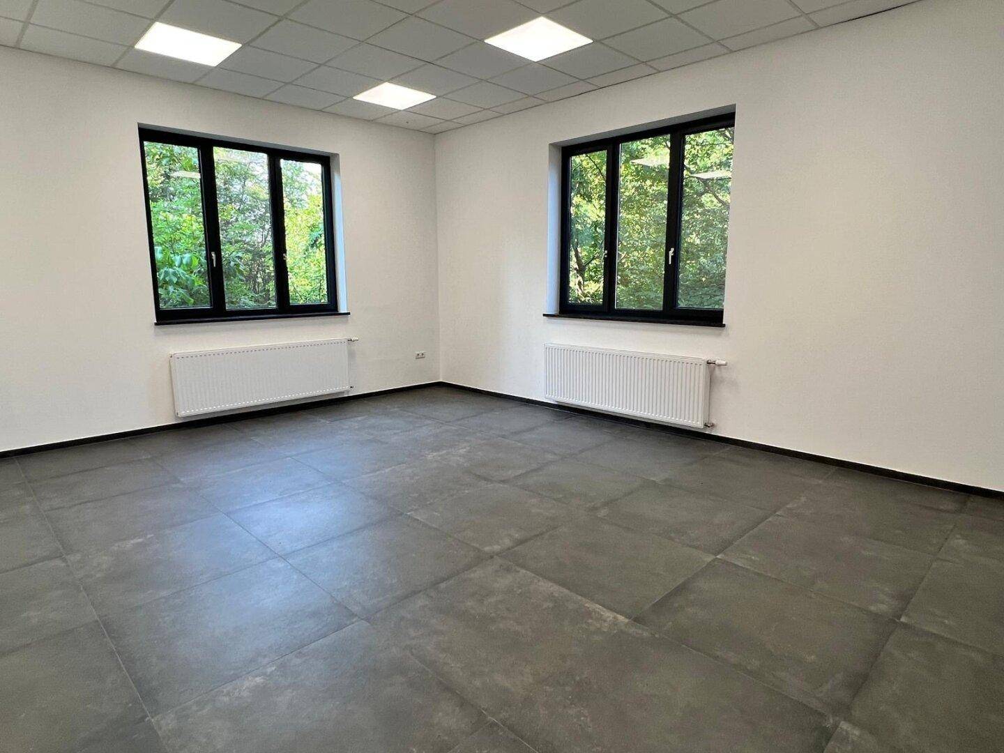 Heller Büroraum mit dunklem Fliesenboden, zwei großen Fenstern und Heizkörpern, die einen grünen Ausblick bieten.
