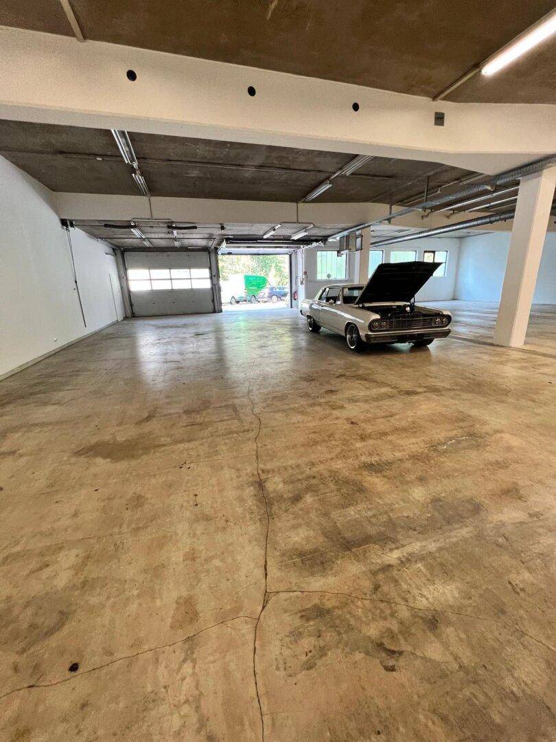 Weitläufige Industriehalle mit Betonboden, hohen Decken und einem klassischen Auto mit geöffneter Motorhaube.
