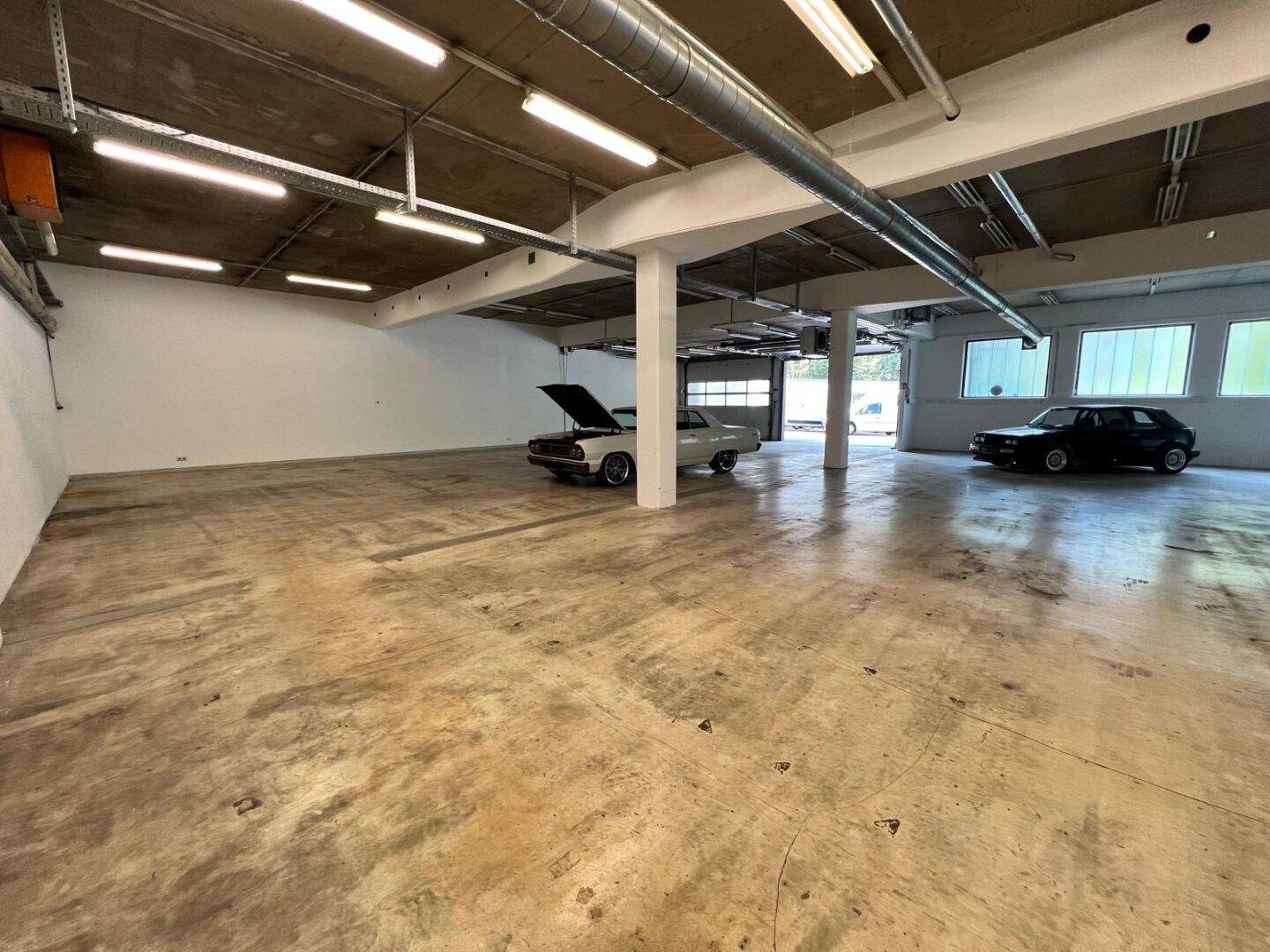 Große Industriehalle mit Betonboden, sichtbaren Rohren und zwei klassischen Autos, die im hinteren Bereich stehen.