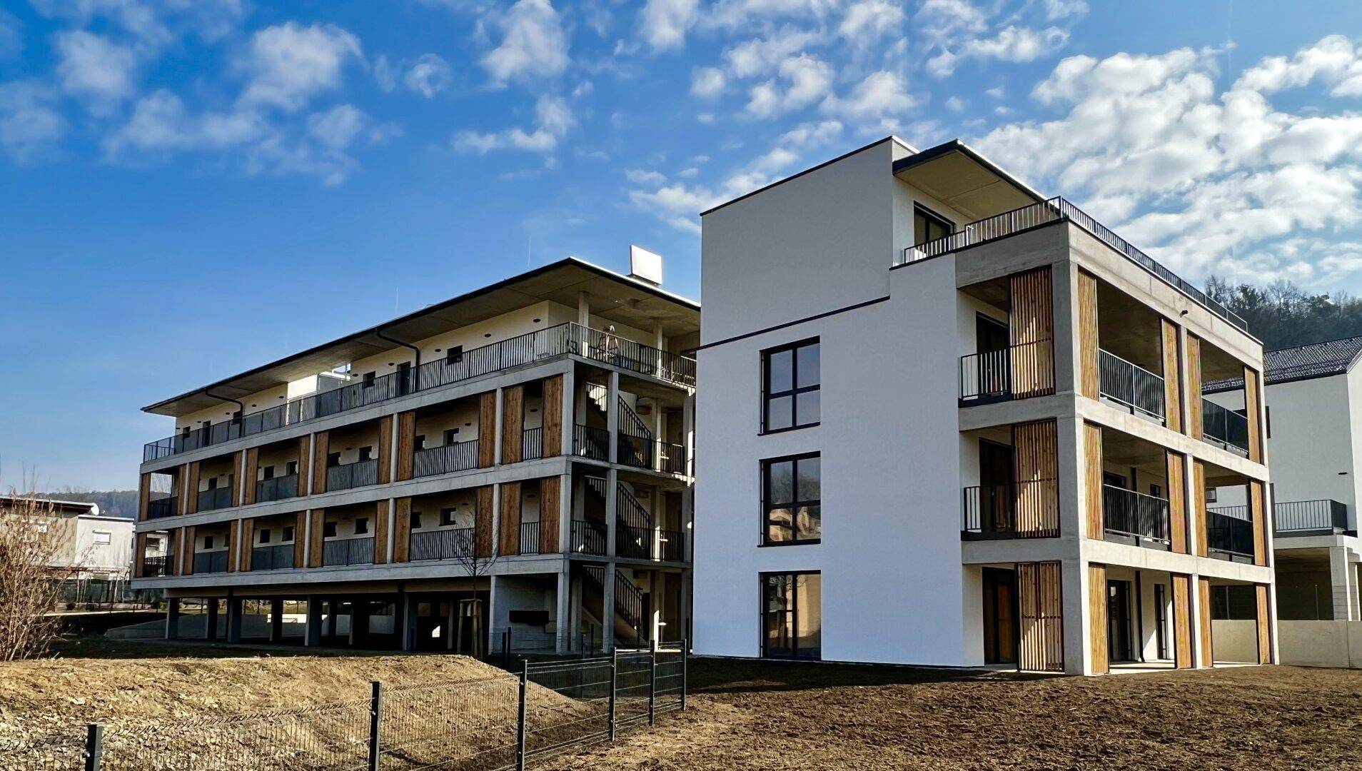 Moderne Mehrfamilienhäuser mit klaren Linien, Beton- und Holzelementen unter blauem Himmel.