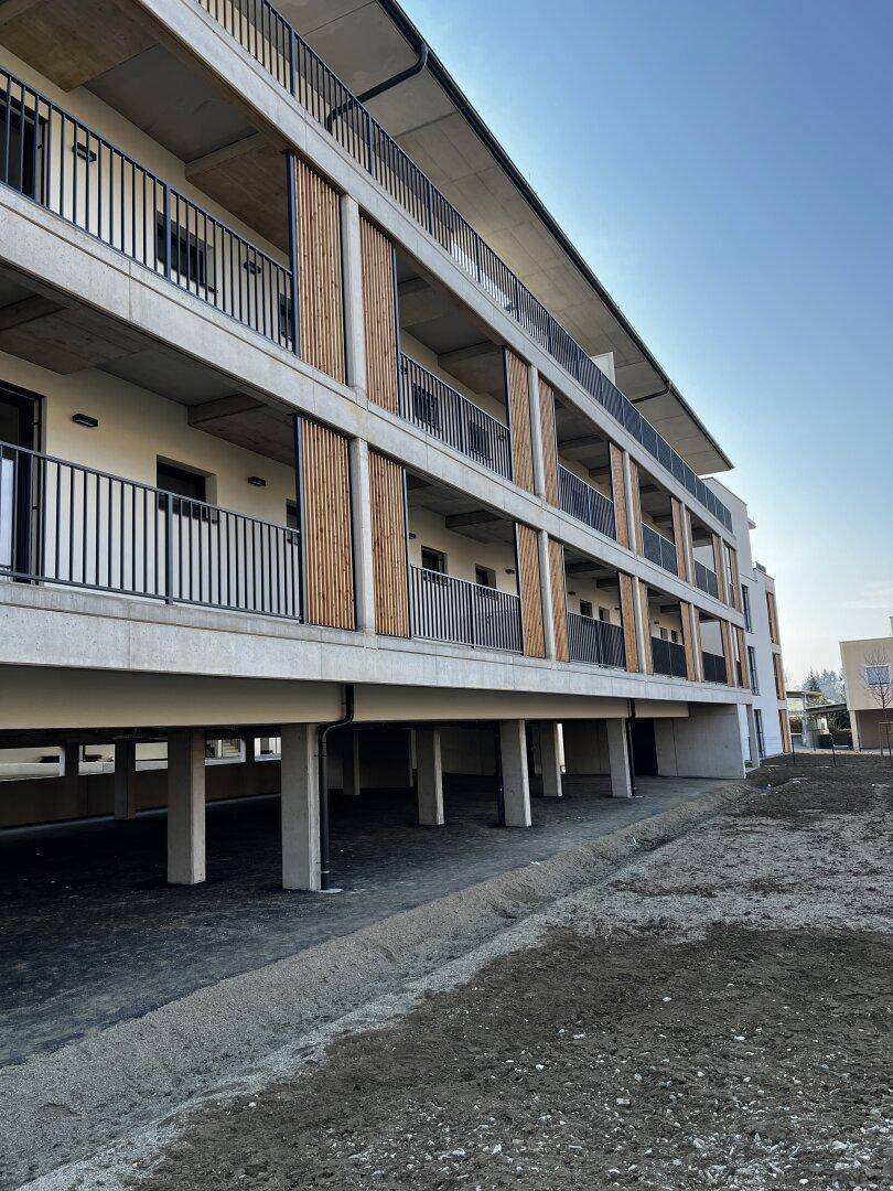 Moderne Wohnanlage mit offenem Parkbereich im Erdgeschoss und Balkonen mit Holzverkleidung darüber.