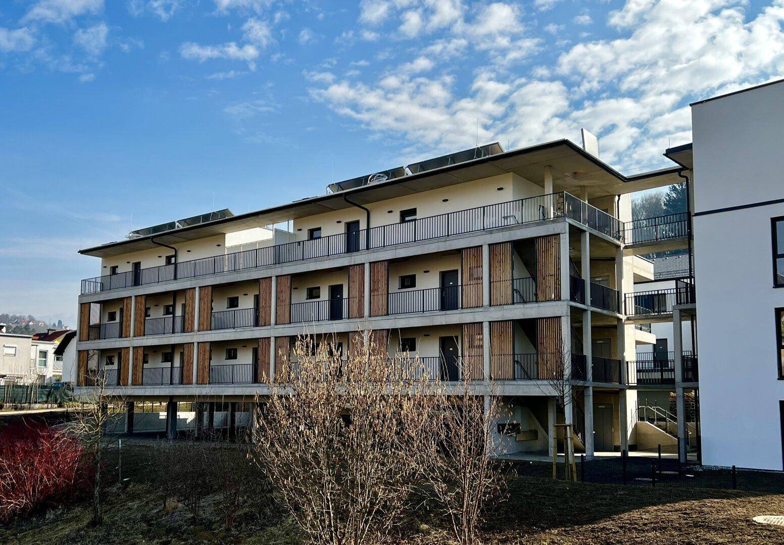Gesamtansicht des modernen Mehrfamilienhauses mit Holzverkleidung, Balkonen und Solaranlagen auf dem Dach.