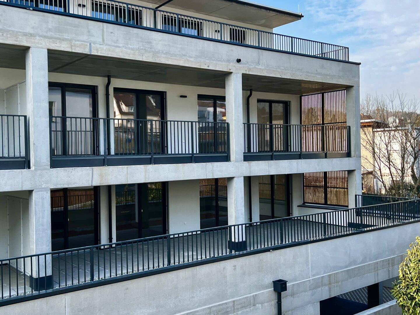 Moderne Wohnanlage mit mehreren Etagen, Betonfassade und schwarzen Balkongeländern unter blauem Himmel.