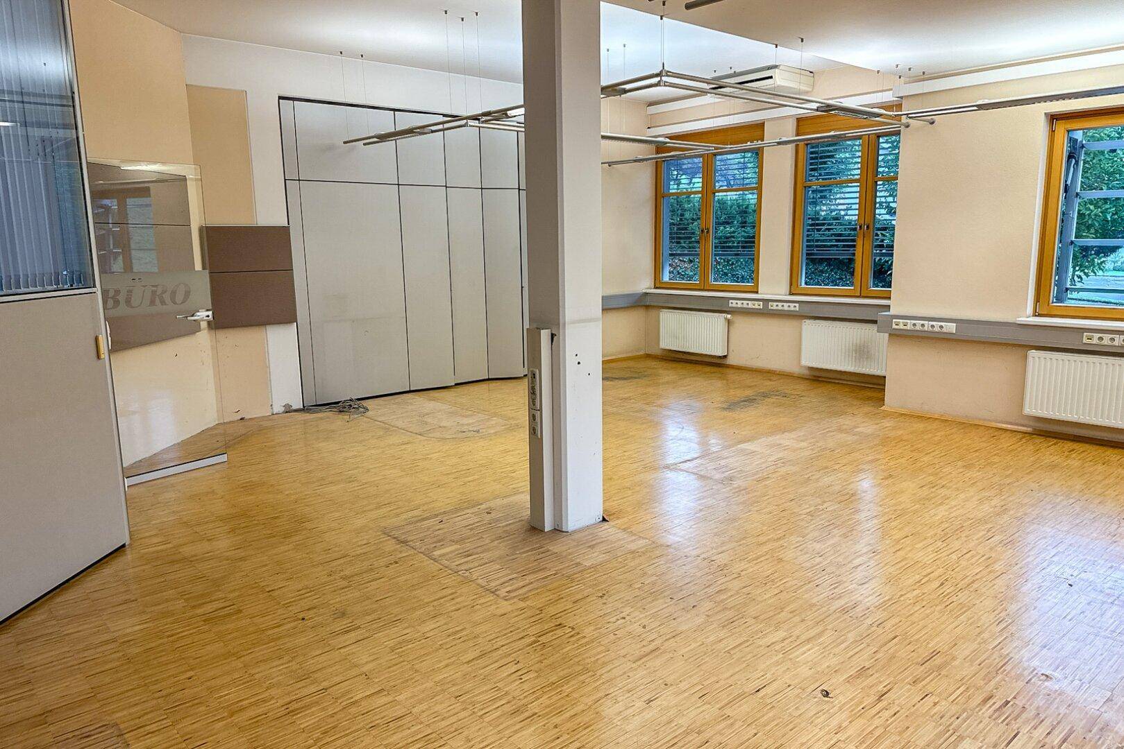 Geräumiger, leerer Büroraum mit Holzparkettboden, vielen Fenstern und einem tragenden Pfeiler.