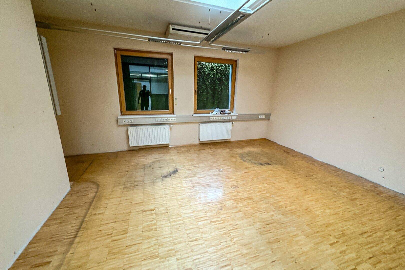 Kleinerer, leerer Büroraum mit Holzparkettboden und zwei Fenstern, die für ausreichend Helligkeit sorgen.