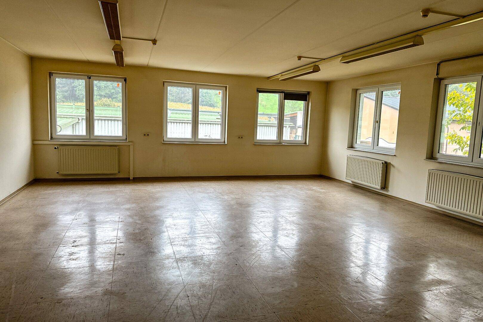 Großer, leerer Raum mit beigefarbenen Wänden, Linoleumboden und vielen Fenstern, die für Helligkeit sorgen.