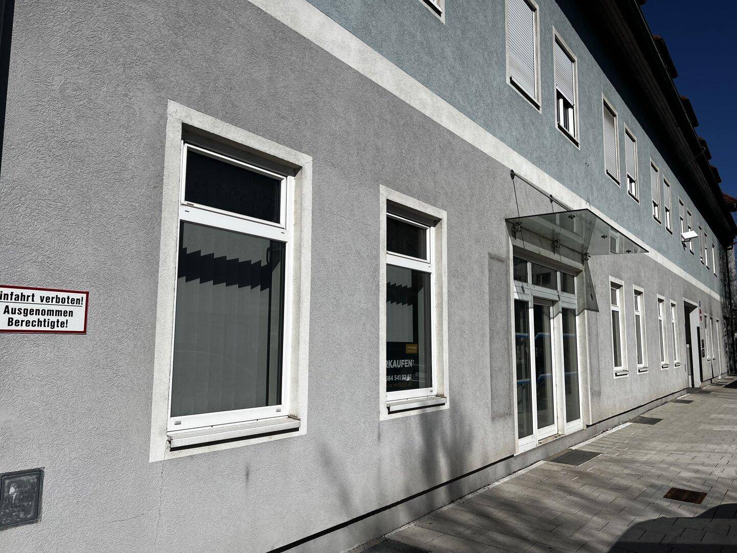 Seitenansicht eines Gebäudes mit grauer Fassade, weißen Fenstern und einem Eingangsbereich.
