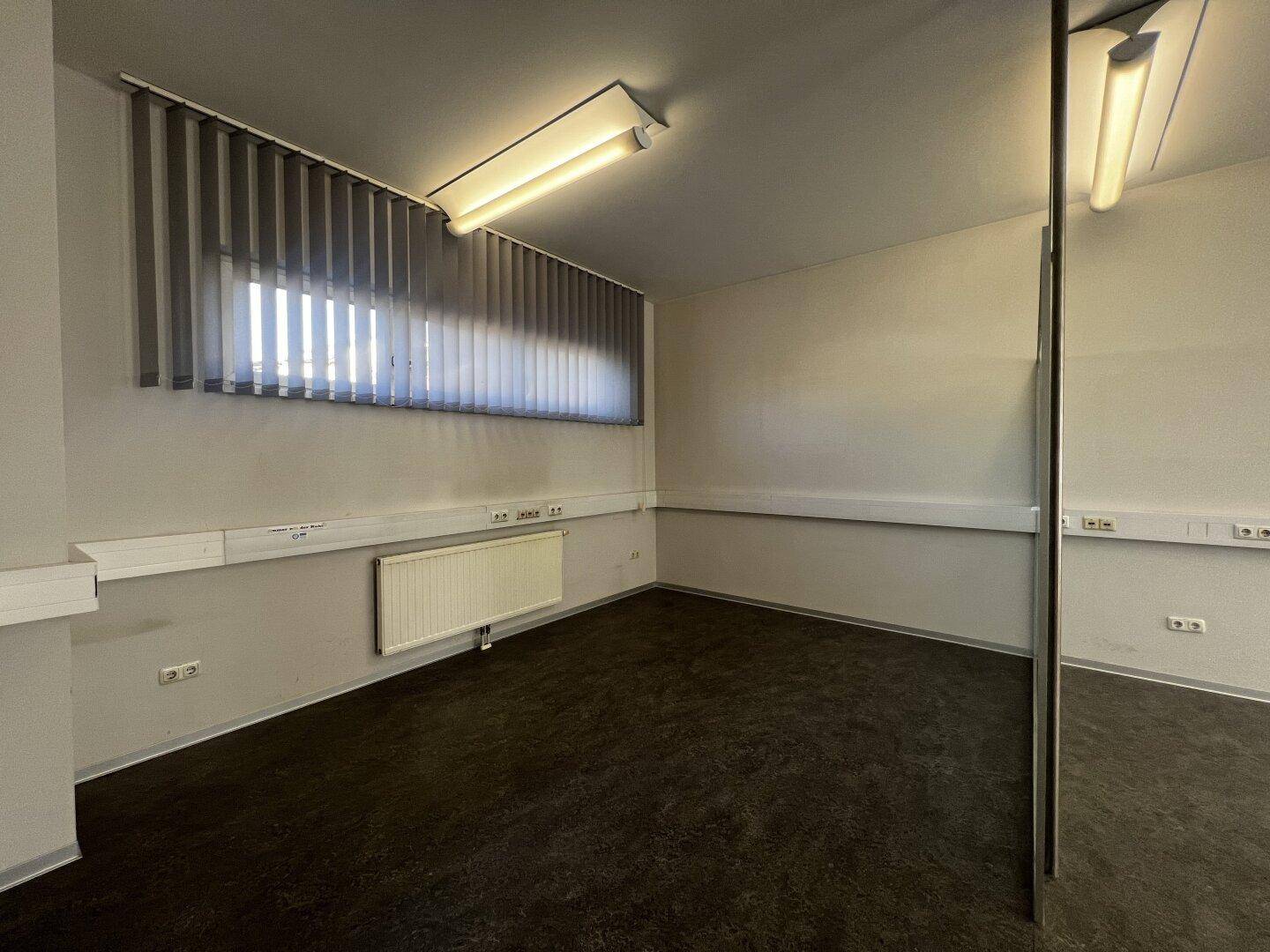 Leerer Büroraum mit dunklem Boden, hellen Wänden und einem breiten Fenster mit Jalousien.