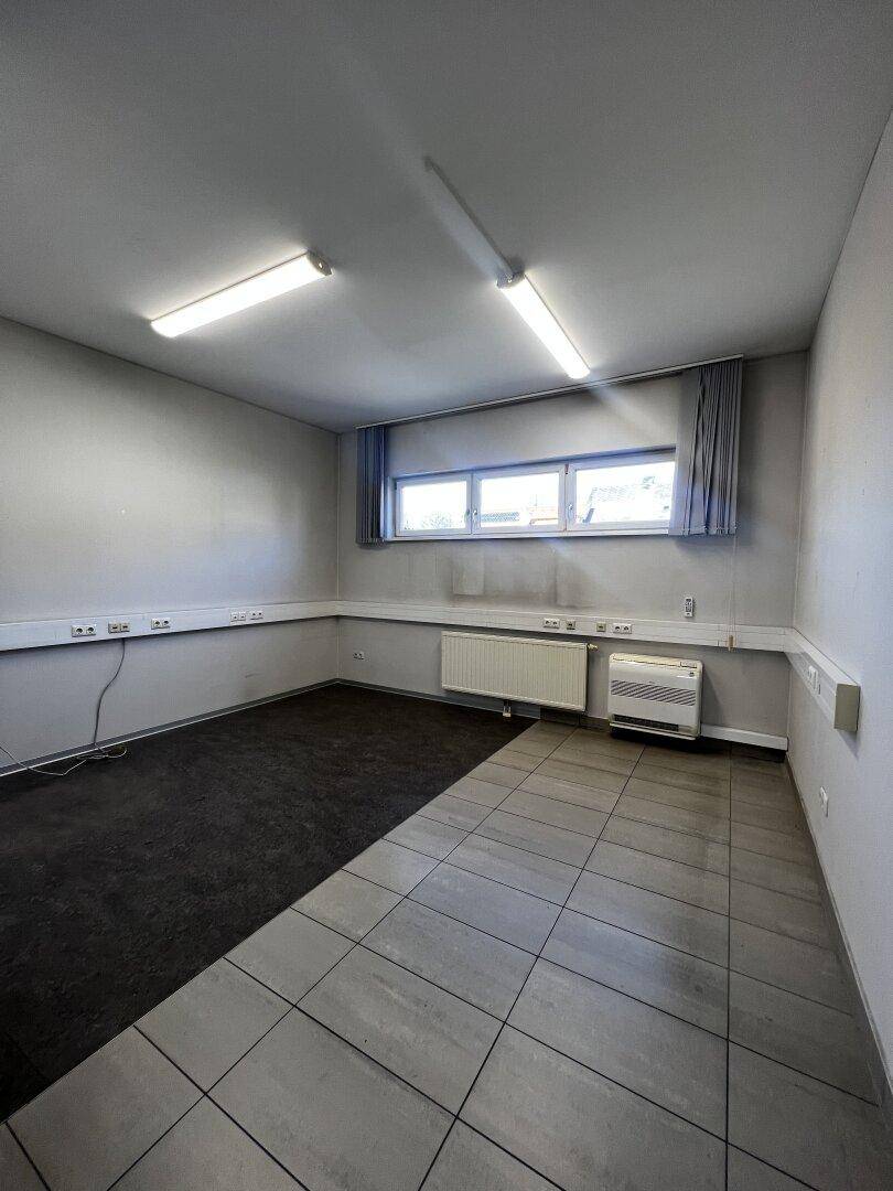 Großer Büroraum mit geteiltem Bodenbelag aus Fliesen und Teppich, sowie einem Fensterband.