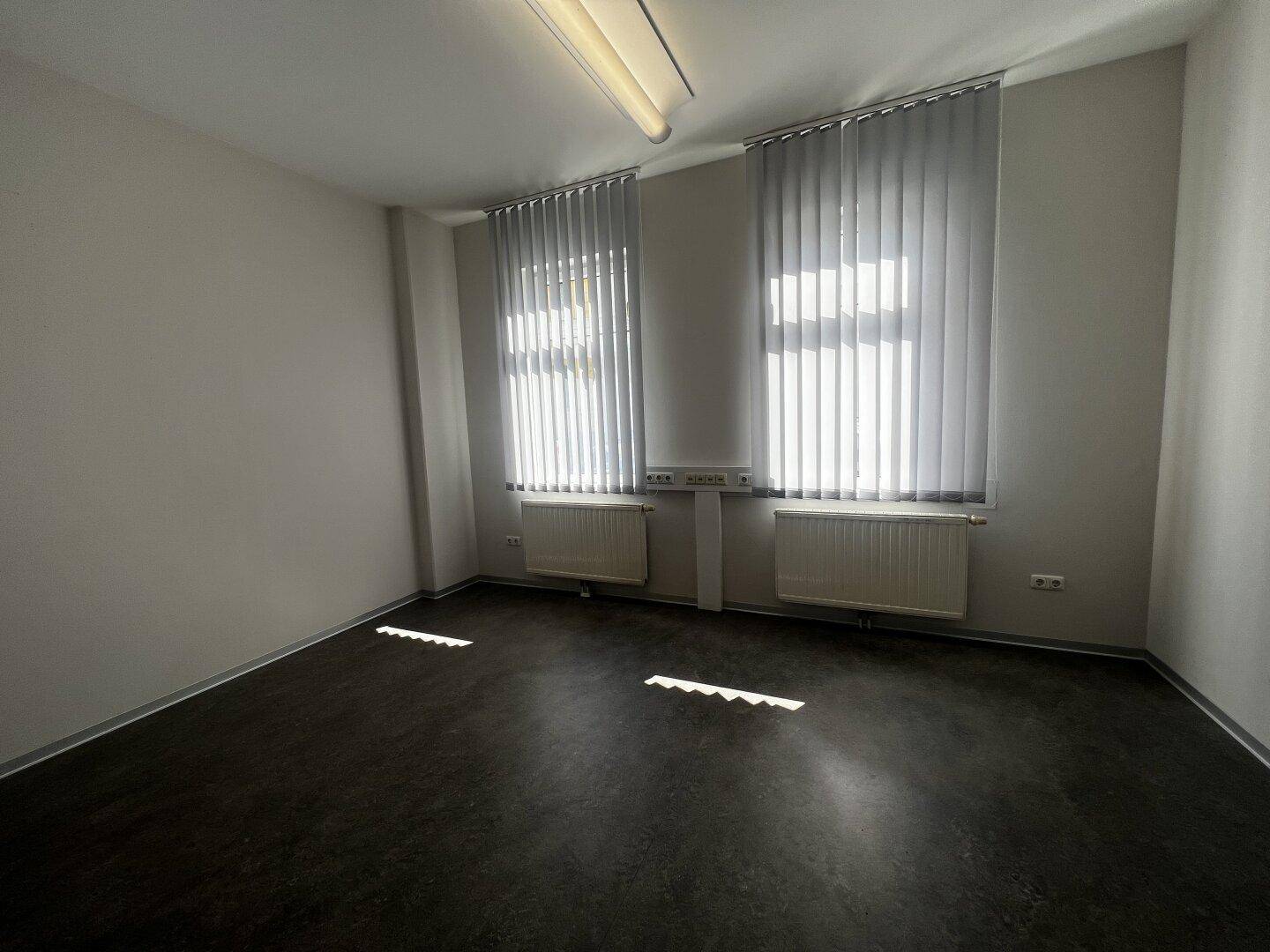 Leerer Büroraum mit dunklem Boden, hellen Wänden und vertikalen Jalousien an den beiden Fenstern.
