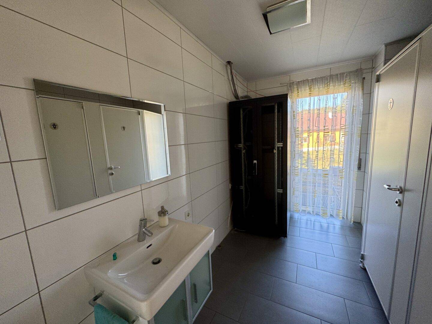 Badezimmer mit weißen Fliesen, Waschbecken, Spiegelschrank und Fenster für Tageslicht.